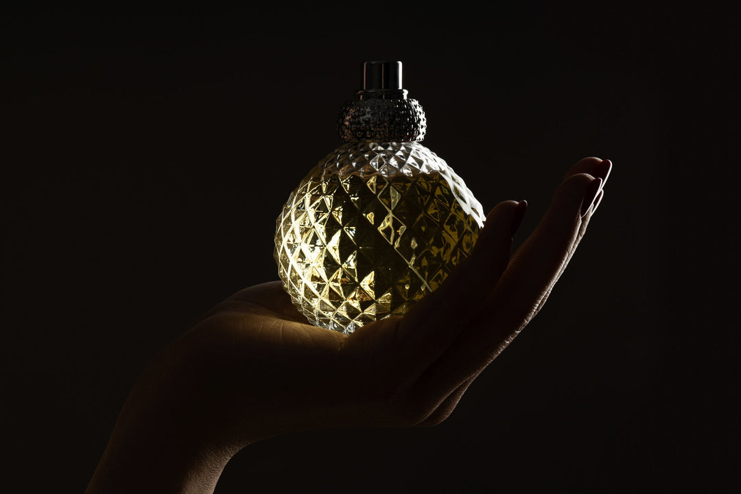 A faceted glass bottle of deep amber fragrance cradled in a cupped hand, lit from one side against a dark background.