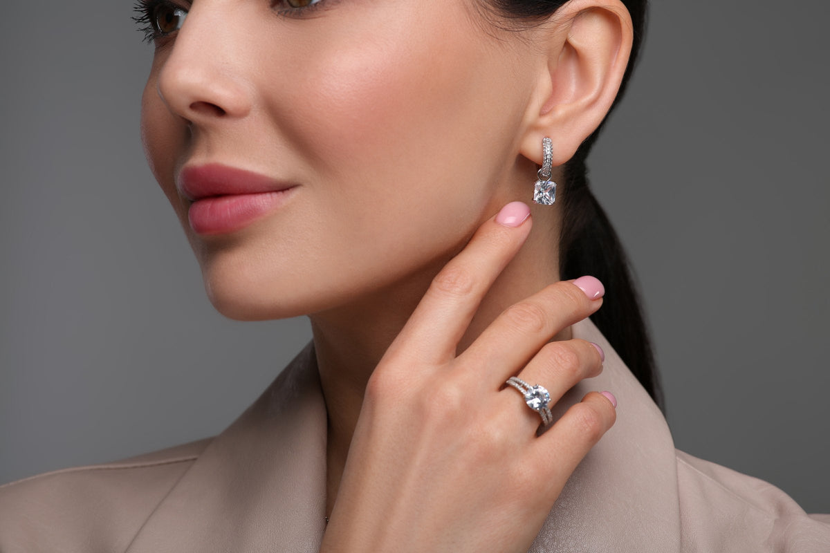 A woman in profile against a dark grey background, wearing a single crystal drop earring and a solitaire ring