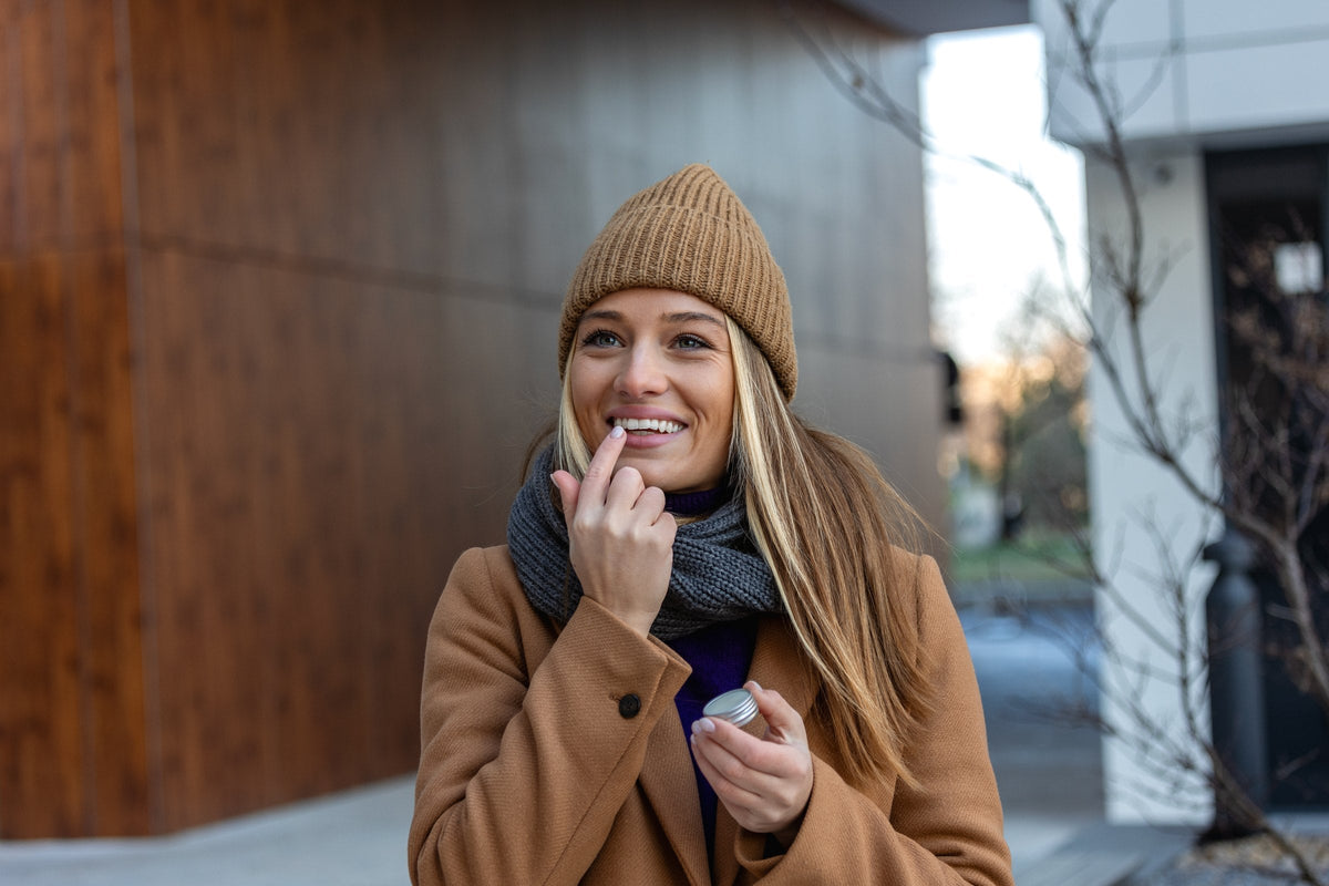 A smiling woman applying lip balm outdoors on a cold day